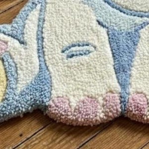 May include: A handmade rug in the shape of a bunny's bottom, featuring a fluffy texture. The rug has a light blue, white, yellow, and pink color scheme. A wooden tag with the text "SAQUITA Rug Decor" is attached.