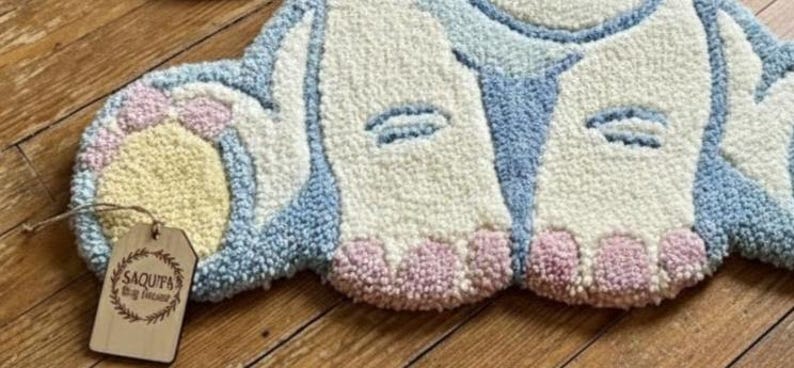 May include: A handmade rug in the shape of a bunny's bottom, featuring a fluffy texture. The rug has a light blue, white, yellow, and pink color scheme. A wooden tag with the text "SAQUITA Rug Decor" is attached.