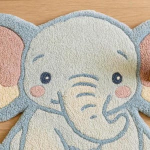 May include: A light blue, ivory, and pink elephant-shaped rug. The rug features a smiling elephant face with blue eyes, pink cheeks, and large ears. The rug is made of a soft, textured material, perfect for a nursery or playroom.