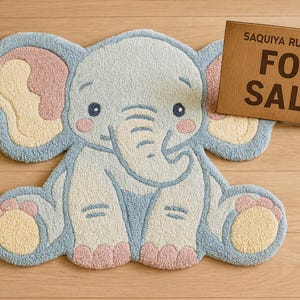 May include: A light blue elephant-shaped rug with pink and yellow accents. The rug has a cartoon-like design and is made of a soft, textured material. A wooden sign that says "FOR SALE" is next to the rug.
