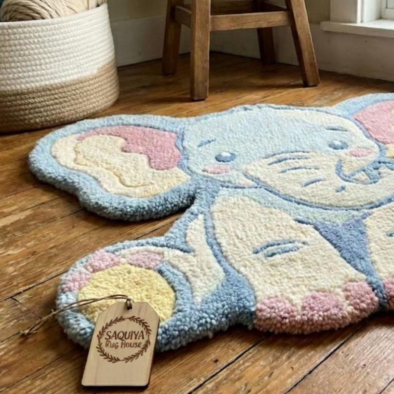 May include: A light blue, ivory, pink, and yellow elephant-shaped rug. The rug has a tufted texture and a cartoon-like design. A wooden tag with the text "SAQUIYA Rug House" is attached to the rug.