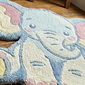 May include: A soft, tufted rug in the shape of an elephant. The rug features a light blue body, pink ears, and accents of yellow and cream. The elephant's face has embroidered details, including eyes and a trunk. This decorative rug is designed for a child's room.