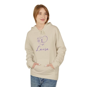 Könnte beinhalten: Beigefarbener Kapuzenpullover mit einem lila herzförmigen Design mit einem Schneeflockenmuster, dem Buchstaben "L" und dem Namen "Laura". Der Hoodie hat eine Kapuze mit Kordelzug, eine Vordertasche und lange Ärmel. Die Person trägt blaue Jeans.