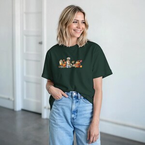 Könnte beinhalten: Dunkelgrünes T-Shirt mit einer Aquarellillustration eines Golden Retrievers, Kürbissen und Herbstlaub. Die Grafik befindet sich auf der Brust. Das T-Shirt wird mit einer blauen Jeans getragen.