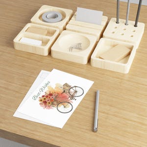 Könnte beinhalten: Ein hölzerner Schreibtisch-Organizer mit mehreren Fächern, darunter ein Klebebandabroller, ein Stifthalter und kleine Ablagen. Eine Postkarte mit einer Illustration eines Fahrrades mit Blumen und dem Text "Best Wishes" liegt auf dem Schreibtisch, zusammen mit einem silbernen Stift.