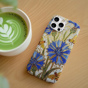 Könnte beinhalten: Smartphone-Hülle mit floralem Design mit blauen Blumen, goldenen Akzenten und Bienen. Eine Tasse grüner Matcha Latte mit einem herzförmigen Design befindet sich neben der Handyhülle. Der Hintergrund ist eine helle Holzoberfläche.