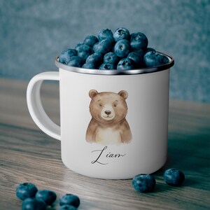 Könnte beinhalten: Weißer Emaillebecher mit einer Aquarellillustration eines braunen Bären und dem Namen "Liam" in Schreibschrift. Der Becher ist mit frischen Blaubeeren gefüllt und steht auf einer Holzoberfläche.