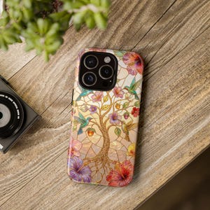 Könnte beinhalten: Bunte Handyhülle mit einem Buntglasdesign, das einen Baum, Kolibris und Blumen in Rosa, Lila und Grün zeigt. Die Hülle liegt auf einer Holzoberfläche.