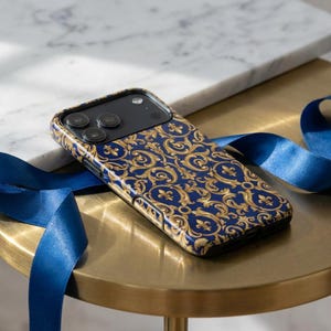 Könnte beinhalten: Eine Smartphone-Hülle mit marineblauem Hintergrund und einem goldenen Barockmuster. Die Hülle liegt auf einem goldenen Tisch mit einem blauen Band. Die Handyhülle hat ein schwarzes Kameramodul.