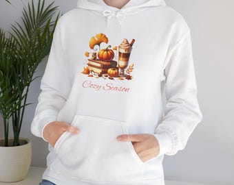 Könnte beinhalten: Weißer Kapuzenpullover mit einer Vordertasche und einem Kordelzug. Auf der Vorderseite befindet sich eine Herbst-Grafik mit Kürbissen, Büchern, Blättern und einem Latte. Der Text "Cozy Season" ist darunter gedruckt.
