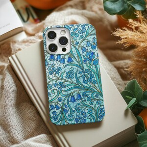Könnte beinhalten: Eine Smartphone-Hülle mit floralem Design in Blau- und Türkistönen, mit glockenförmigen Blumen und aufwendigen Mustern. Die Hülle liegt auf einem geschlossenen Buch mit beigem Einband, vor einem weichen Hintergrund.