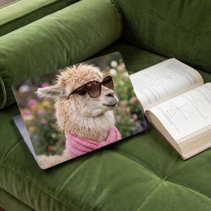 Op de afbeelding: Laptop met een hoes met een lichtbruine lama met een zonnebril en een roze sjaal. De lama staat tegen een vage bloemenachtergrond. De laptop is open, naast een open boek op een groene fluwelen bank.