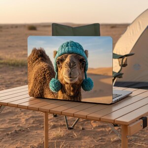 Könnte beinhalten: Ein Laptop mit einem Kamel mit blauer Strickmütze und Bommeln steht auf einem klappbaren Holztisch. Ein Campingzelt und ein Stuhl befinden sich im Hintergrund, vor einer Wüstenlandschaft.