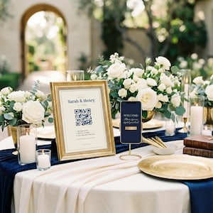 Könnte beinhalten: Ein goldgerahmtes Schild mit dem Text Sarah & Henry, einem QR-Code und Anweisungen, neben einem Smartphone auf einem Goldständer, weißen Rosen, Kerzen und einem marineblauen Tischläufer auf einer cremefarbenen Tischdecke.