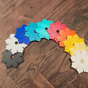 Könnte beinhalten: Eine Sammlung farbenfroher, sternförmiger Untersetzer, die in einem Bogen angeordnet sind. Die Untersetzer sind in verschiedenen Farben gehalten, darunter Grau, Weiß, Blau, Türkis, Rosa, Orange und Gelb. Jeder Untersetzer hat ein kreisförmiges Loch in der Mitte.