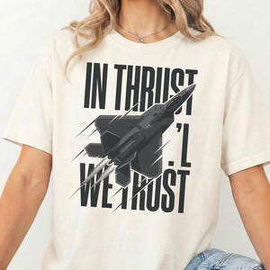 Puede incluir: Camiseta color crema con un gráfico de avión negro y el texto "IN THRUST, I TRUST". El diseño incluye líneas de velocidad estilizadas. La camiseta es de corte holgado.