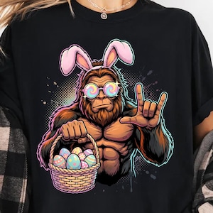 May include: Black t-shirt featuring a cartoon Bigfoot wearing bunny ears and sunglasses, holding a basket of Easter eggs. The Bigfoot is making a rock-on hand gesture. The design has a colorful, graphic style.
