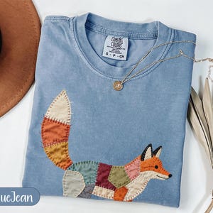May include: Blue jean-colored t-shirt with a patchwork fox design. The fox is made of various colors, including orange, red, green, and brown. The shirt has a crew neck and short sleeves. A gold necklace with a pendant is draped across the shirt.