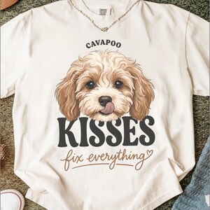 May include: An off-white short-sleeve t-shirt featuring a graphic of a curly-haired Cavapoo dog with its tongue out. The text above reads CAVAPOO and below the dog it says KISSES fix everything in a mix of bold black and brown script fonts.