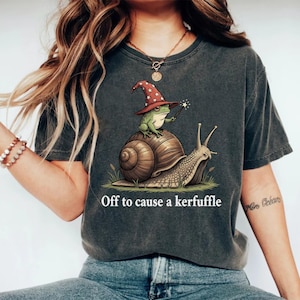 May include: Dark gray t-shirt featuring a whimsical illustration of a frog wearing a wizard hat and riding a snail. The text "Off to cause a kerfuffle" is printed below the image. The design is printed on a soft, comfortable tee.