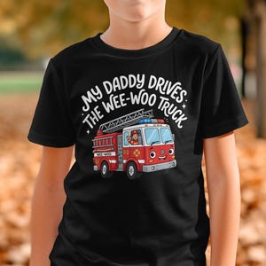 Può includere: Una t-shirt nera a maniche corte con la grafica di un camion dei pompieri rosso e bianco con un volto da cartone animato. Sopra il camion, il testo bianco recita: MY DADDY DRIVES THE WEE-WOO TRUCK. Il camion ha una scala, luci e un conducente.