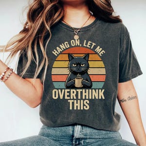 May include: Dark grey t-shirt with a graphic of a black cat holding a mug, set against a retro sunset design. The text reads "Hang on, let me overthink this."