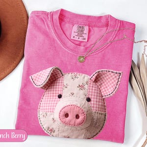 May include: A pink Comfort Colors t-shirt featuring a patchwork pig face design. The pig's face is made of various fabrics, including gingham and floral patterns. A gold necklace with a pendant rests on the shirt. The text "Crunch Berry" is in a pink oval.