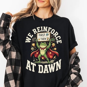 Op de afbeelding: Een zwart t-shirt met een afbeelding van een groene kikker in een rode cape en vlinderdas. De kikker houdt een bordje vast met "FIRST THEN" en iconen van een boek en trofee. De tekst "WE REINFORCE AT DAWN" staat in een boog om de afbeelding.