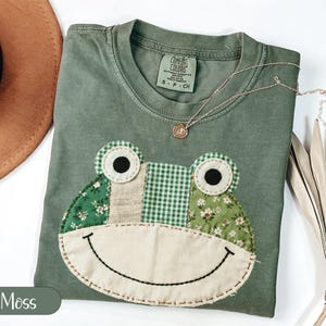 May include: A moss green t-shirt with a frog face applique. The frog face is made of various green patterned fabrics, including floral and gingham. The shirt has a round neckline and short sleeves. A gold necklace with a small pendant is on the shirt.
