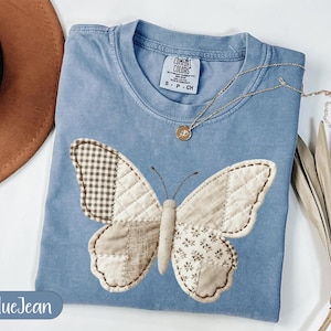 Könnte beinhalten: Ein blaues Jeans-T-Shirt mit einem großen Schmetterlings-Applikation. Der Schmetterling hat Patchwork-Flügel in verschiedenen neutralen Tönen und ein Steppmuster. Eine goldene Halskette mit einem kleinen Anhänger liegt auf dem Shirt. Das Shirt hat den Text "BlueJean".