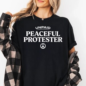 Black t-shirt with white text "UNPAID PEACEFUL PROTESTER" and a peace symbol.