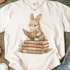May include: A white t-shirt with a whimsical design of a bunny wearing spectacles and a bow tie, reading a book atop a pile of books. The illustration has a vintage feel. The shirt has the text "Comfort Colors" on the collar.