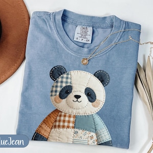 May include: A blue jean colored t-shirt with a patchwork panda bear design. The panda has blue, white, and brown patches. A gold necklace with a small pendant is draped across the shirt. The shirt has a Comfort Colors tag.