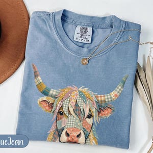 May include: Blue jean colored t-shirt with a colorful patchwork highland cow graphic. The cow's horns and face are made of various patterned fabrics. A gold necklace with a small pendant rests on the shirt. The shirt is labeled "Comfort Colors".