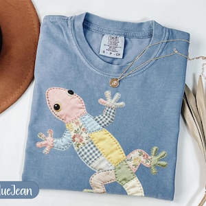 May include: A blue jean colored t-shirt with a patchwork lizard design. The lizard is made of various fabric patterns and colors, including pink, yellow, and floral prints. A gold necklace with a pendant rests on the shirt.