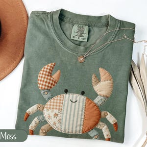 May include: Moss green t-shirt with a patchwork crab design. The crab features various patterned fabrics in orange, brown, and blue. A gold necklace with a small pendant rests on the shirt. The shirt is folded, with a brown hat and dried palm leaves in the background.