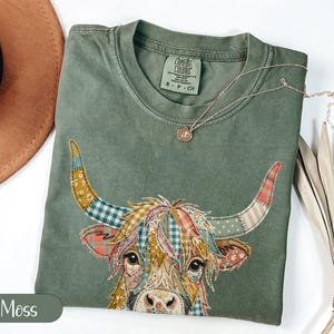 Puede incluir: Camiseta verde musgo con un diseño de retazos de colores de la cabeza de una vaca de las Tierras Altas. Los cuernos y la cara de la vaca están adornados con varias telas estampadas. Un delicado collar descansa sobre la camisa. Un sombrero marrón en el fondo.