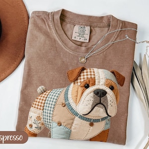 May include: Espresso-colored t-shirt with a patchwork bulldog design. The bulldog features brown, tan, blue, and white fabric patches. A silver necklace with a small pendant rests on the shirt. The shirt is folded, with a brown hat visible in the background.