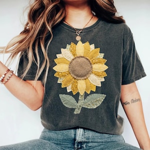 Pode incluir: T-shirt cinza escuro com um grande design de girassol aplicado. O girassol tem pétalas amarelas e creme, um centro de serapilheira marrom e folhas verdes. A camisa é de estilo casual.