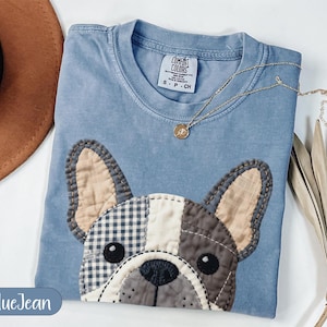 May include: A blue jean colored t-shirt with a patchwork French bulldog design. The dog's face features various fabric patterns and colors, including gingham and solid gray. A gold necklace with a small pendant rests on the shirt.