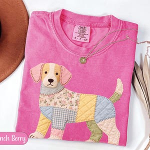 Peut inclure: Un t-shirt rose Comfort Colors avec un motif de chien en patchwork. Le chien a une tête beige, des oreilles roses et un corps fait de divers tissus à motifs. Un collier en or avec un pendentif est drapé sur le t-shirt. Le texte "Crunch Berry" est dans un ovale rose.
