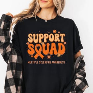 May include: Black t-shirt with the words "SUPPORT SQUAD" in orange, with a matching orange ribbon and floral accents. The text "MULTIPLE SCLEROSIS AWARENESS" is below the main text. The shirt is worn by a person.