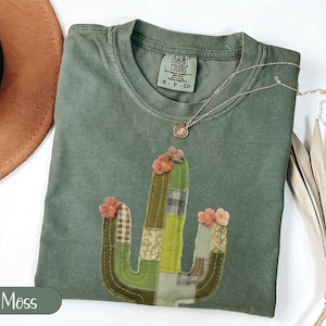 May include: A moss green t-shirt with a cactus applique. The cactus is made of various green and tan fabrics and adorned with small pink flowers. A gold necklace with a pendant rests on the shirt. The shirt is folded next to a brown hat.