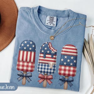 Puede incluir: Camiseta azul claro con diseño patriótico de paletas heladas. Las paletas presentan patrones rojos, blancos y azules, incluyendo estrellas y rayas, con lazos en la parte inferior. La camiseta tiene cuello redondo y una etiqueta pequeña.