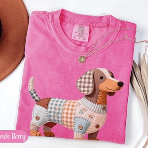 May include: A pink Comfort Colors t-shirt featuring a patchwork dachshund design. The dog has a brown and tan body with various patterned patches and button accents. A gold necklace with a pendant is draped across the shirt. The text "Crunch Berry" is in a pink bubble.
