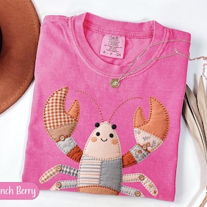 Puede incluir: Una camiseta rosa brillante Comfort Colors con un diseño de langosta de patchwork. La langosta tiene pinzas y segmentos corporales estampados, con una cara sonriente. Un collar de oro con un pequeño colgante descansa sobre la camisa. El texto "Crunch Berry" está en un óvalo rosa.