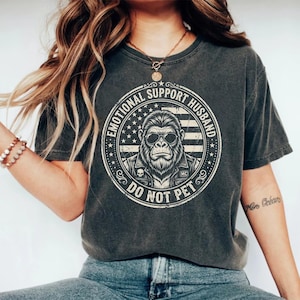 May include: Dark gray t-shirt featuring a circular graphic with a gorilla wearing sunglasses and a leather jacket, set against an American flag design. The text reads "EMOTIONAL SUPPORT HUSBAND" and "DO NOT PET".
