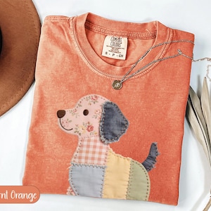 May include: Burnt orange t-shirt with a patchwork dog design. The dog has a floral patterned ear, a gingham body, and a blue tail. A silver necklace with a small pendant is on the shirt. The shirt has a Comfort Colors tag.