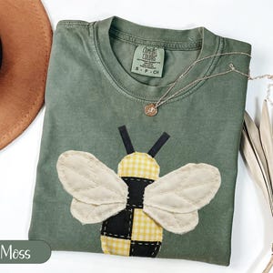 Puede incluir: Una camiseta verde musgo con un gran aplique de abeja. La abeja tiene alas color crema, un cuerpo a cuadros amarillo y negro y antenas negras. Un collar dorado con un pequeño colgante descansa sobre la camisa.