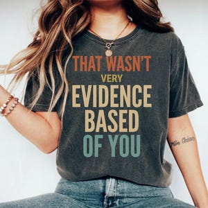 Könnte beinhalten: Dunkelgraues T-Shirt mit dem Text "THAT WASN'T VERY EVIDENCE BASED OF YOU" in Orange, Beige und Türkis. Das T-Shirt hat einen lässigen Stil.
